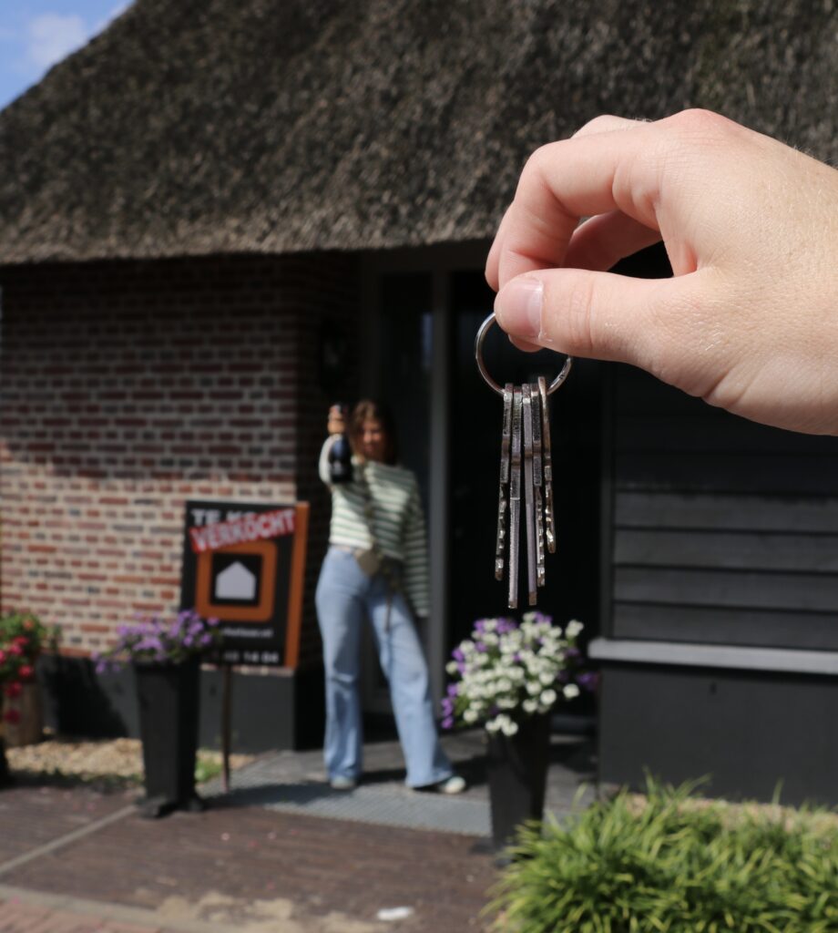 Een hand houdt sleutels omhoog voor een huis met een VERKOCHT-bord en een vrouw die een fles champagne vasthoudt.