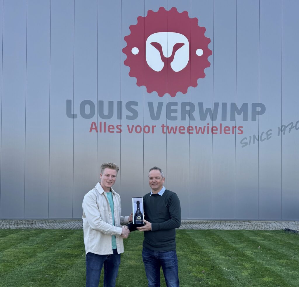 Twee mannen poseren met een fles wijn voor het gebouw van Louis Verwimp met het logo en de slogan Alles voor tweewielers.