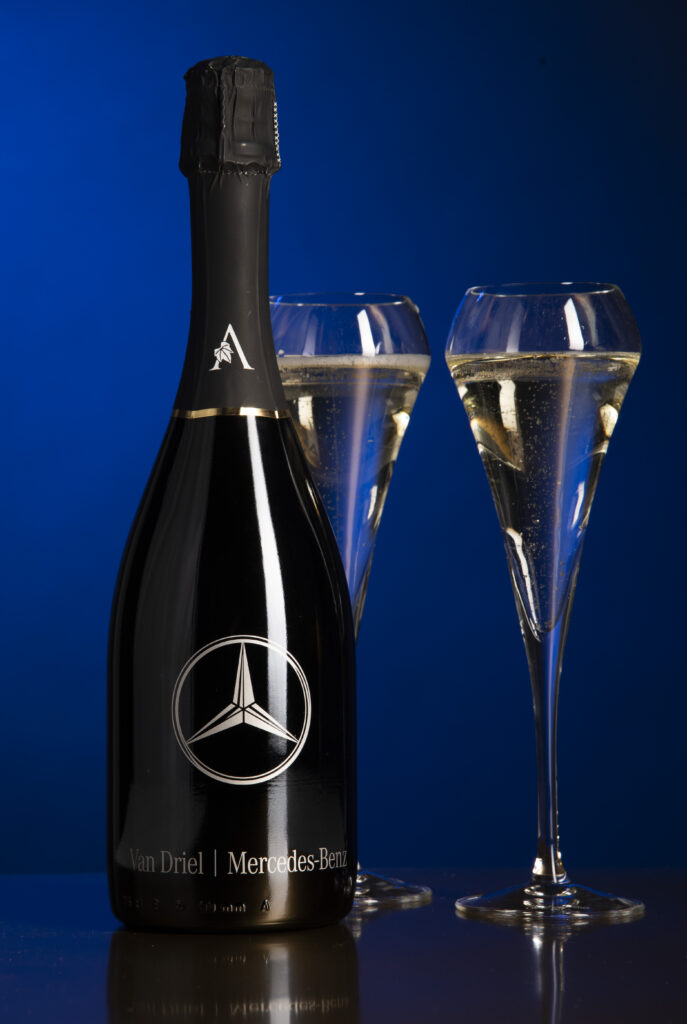 Fles mousserende wijn van Lierno Wines met Mercedes-Benz logo en twee gevulde champagneglazen tegen een blauwe achtergrond.