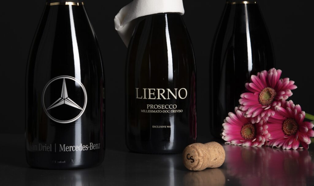 Drie flessen Lierno Prosecco met een Mercedes-Benz logo, een kurk en roze bloemen op een donkere ondergrond.