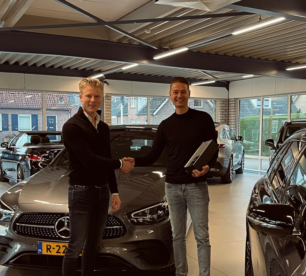 Twee mannen schudden elkaar de hand bij de aflevering van een Mercedes in een autoshowroom.