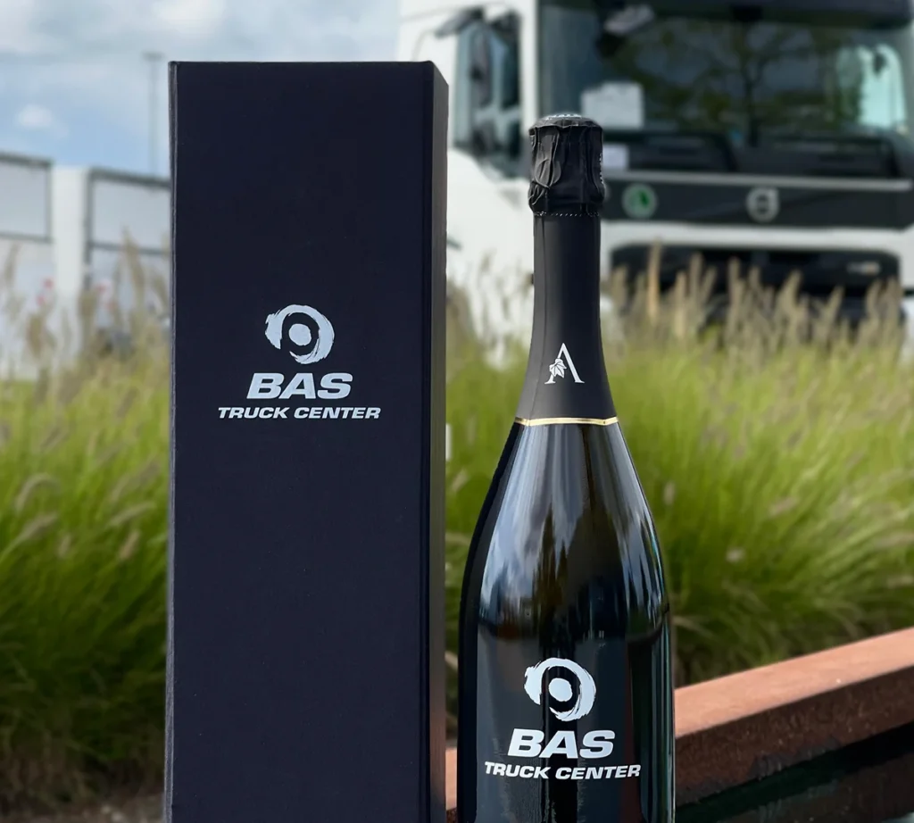 Fles wijn en geschenkdoos met BAS Truck Center-logo op de voorgrond en een vrachtwagen op de achtergrond.
