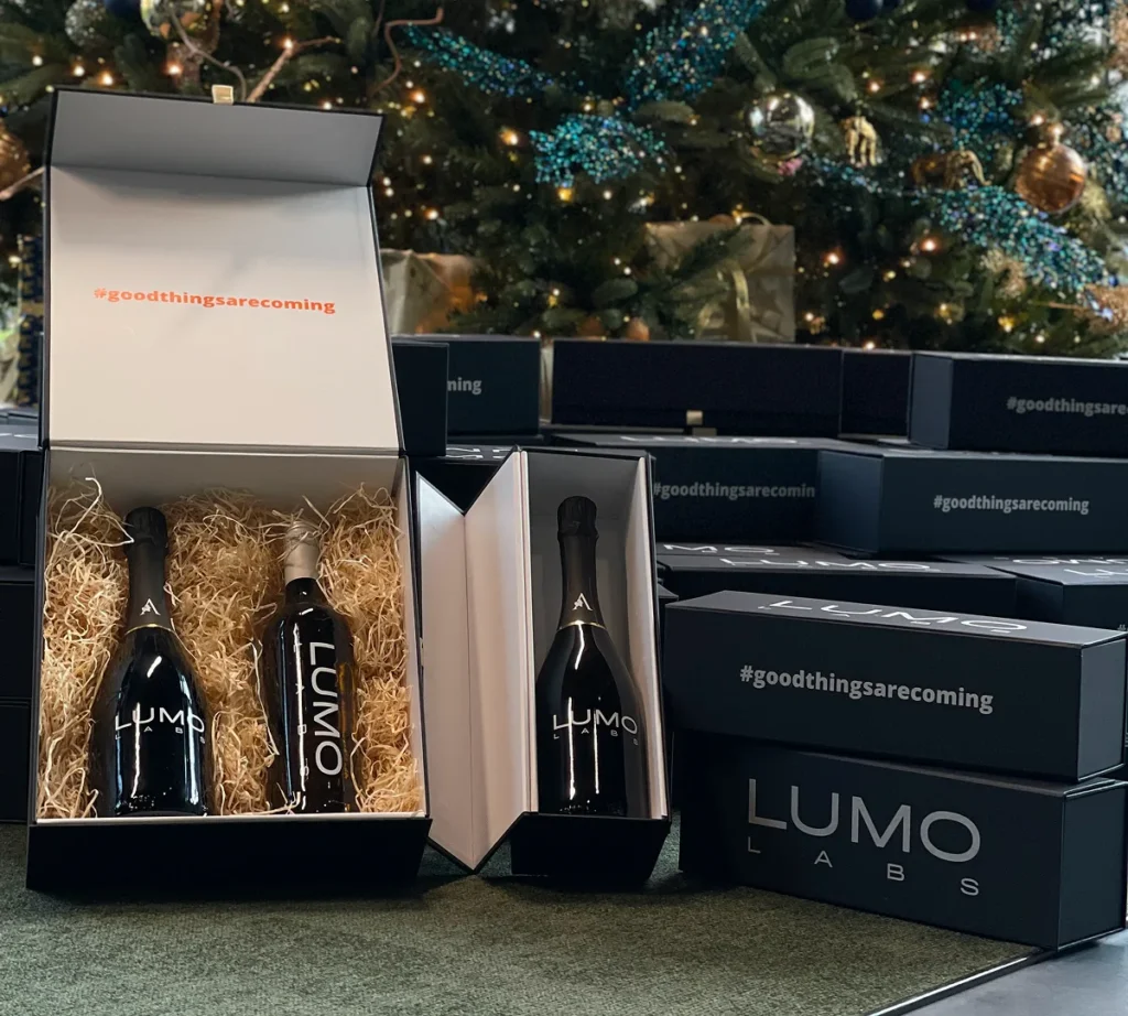 Geschenkverpakkingen met LUMO Labs wijnflessen staan uitgestald voor een kerstboom met versiering.